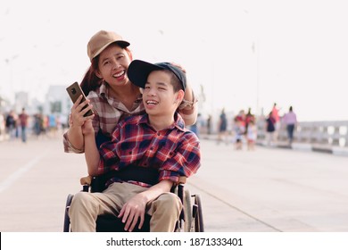 Asian Disabled child on wheelchair and mother fun with selfie by smart phone on the outdoors ​nature​ and seagull birds background​,Lifestyle​ of Happy disability kid travel in family holiday concept.