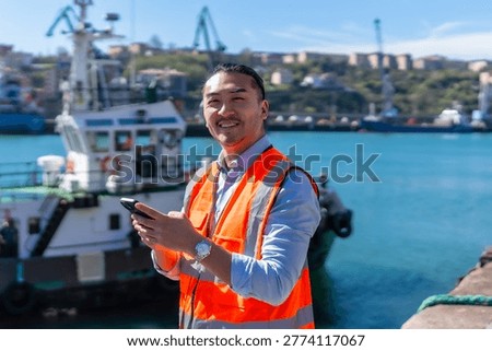 Asian dock worker smiling while standing at a busy port, wearing an orange safety vest and using a smartphone for communication and logistics management