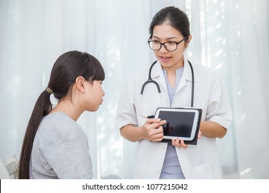 Asian Doctor Having Conversation With Her Asian Teenage Girl Patient And Holding Ipad Gadget In Medical Office. Doctor Intern ( Woman ) Explaining X-ray Results To Patient