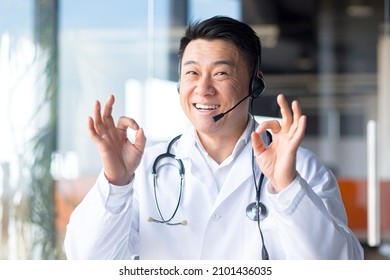  Asian doctor, looking at the camera and having fun talking to the patient, smiling amicably and rejoicing in the patient's recovery, working remotely, video call with headset - Shutterstock ID 2101436035