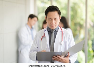 Asian Doctor Man Portrait In Professional Uniform Standing And Holding Folder To Review Data With Smile And Confidence.