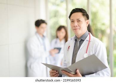 Asian Doctor Man Portrait In Professional Uniform Standing And Holding Folder To Review Data With Smile And Confidence.