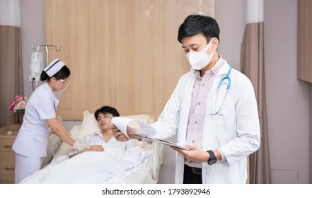 Asian Doctor With Mask Checking Lab Test Results Of Elderly Hospice Patient With Nurse With Face Shield At Hospital. People Health And Medical Concept.