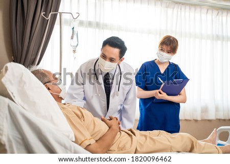 Asian doctor and nurse wearing mask checking health of senior old patient in hospital recovery room. The elder man infected covid lying on bed receiving treatment and diagnosis from medical team.