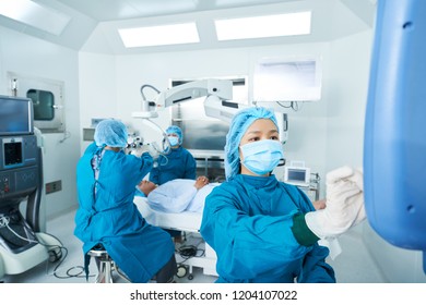 Asian Doctor In Surgeon Uniform Using Modern Monitor While Helping Colleagues To Perform Surgery In Operating Theatre