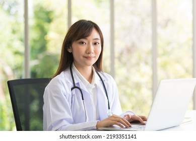 Asian Doctor Woman In White Lab Coat Uniform Sitting And Working With Her Laptop In Modern Office Clinic Room.