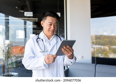 Asian doctor works in a modern clinic, in a private office, conducts an online consultation holding a tablet computer, reports good news, smiles at the camera, happy, video call - Powered by Shutterstock