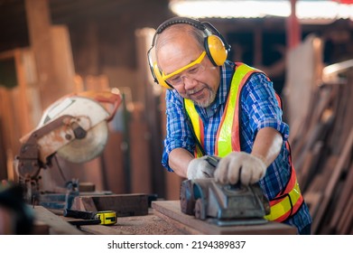 Asian elderly carpenter In workwear safety equipment uniform such as gloves,glasses, ear-muffs or noise-cancel equipment are using an electric planer to smooth out the wood to produce furniture. - Shutterstock ID 2194389837