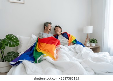 Asian elderly gay couple lying down on bed and holding rainbow flag. Attractive mature senior man LGBTQ people in pajamas enjoy early morning routine activity after wake up together in bedroom at home - Powered by Shutterstock