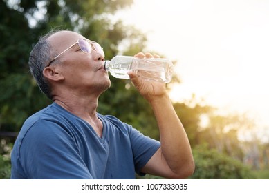 Asian Elderly male in blue shirt with glasses drinking water from bottle after exercise at park outdoor background.