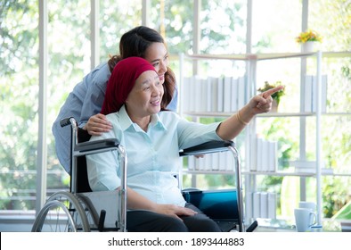 Asian elderly mother who sitting on wheelchair and wearing red headscarf, show her lovely daughter to look at something through window very happily and smile after she survive from cancer or leukemia. - Shutterstock ID 1893444883