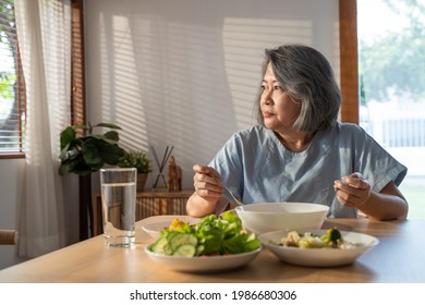 Asian Elderly Retired Grandmother Stay At Home With Painful Face Sitting Alone On Eating Table In House. Depressed Mature Senior Old Woman Upset Feeling Unhappy, Lonely And Missing His Family At Home