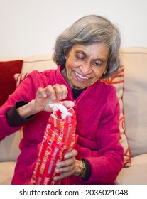 Asian Elderly Woman Opening A Gift Or Present At Christmas. Happy Ethnic Senior Older Lady Sitting At Home And Smiling, UK