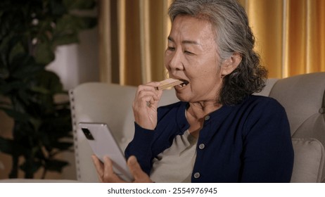 Asian elderly woman sits on sofa, using smartphone in hand and drinking eating snack looking relaxed and engaged with her device. - Powered by Shutterstock