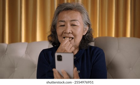 Asian elderly woman sits on sofa, using smartphone in hand and drinking eating snack looking relaxed and engaged with her device. - Powered by Shutterstock