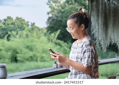 Asian elderly woman is spending her free times at her house balcony by using her mobile phone to do online transaction, and to look after houseplants in small pots nearby, soft focus, new edited. - Powered by Shutterstock