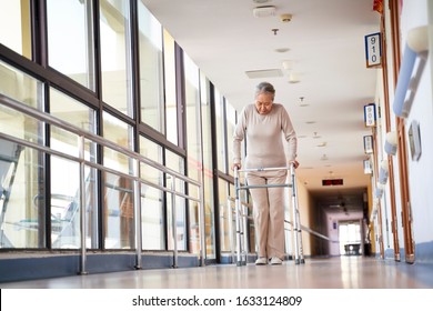 Asian Elderly Woman Walking Using A Walker In Hall Way Of Nursing Home, Low Angle View.