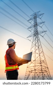 Asian electrical engineers checking location using a notebook computer standing at a power station to view the planning work by producing electrical energy at high voltage electrodes. Vertical image.