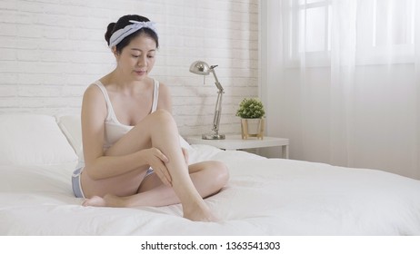 Asian Elegant Woman Caring About Body Legs And Feet Putting Hydrating Cream On It. Young Girl In Shorts Casual Wear Relax Sit On Bed Bright Cozy White Bedroom Near Window Applying Lotion Moisturizer