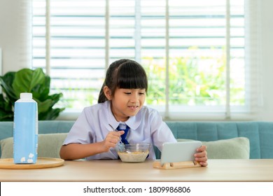 Asian Elementary School Student Girl In Uniform Eating Breakfast Cereals With Milk And Looking Cartoon In Smartphone To Distractions To Eat It In Morning School Routine For Day In Life Getting Ready.