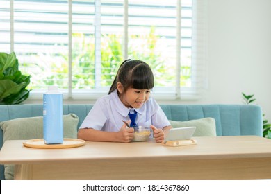 Asian Elementary School Student Girl In Uniform Eating Breakfast Cereals With Milk And Looking Cartoon In Smartphone To Distractions To Eat It In Morning School Routine For Day In Life Ready.