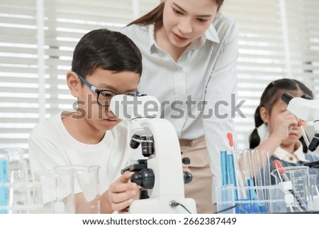 Asian elementary student using microscope during hands-on science class observed by female caucasian teacher standing behind with beakers, test tubes, and blue liquids on lab table