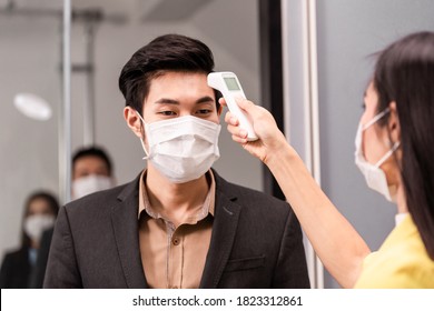 Asian Employee Woman Using Infrared Thermometer Check Fever Temperature Measurement Of Office Worker Wearing Face Mask Before Enter Room At Entrance Door. Coronavirus Prevention, New Normal Working