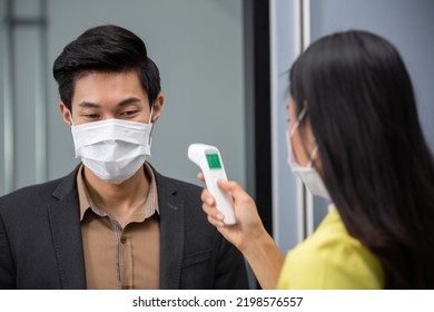 Asian Employee Woman Using Infrared Thermometer Check Fever Temperature Measurement Of Office Worker Wearing Face Mask Before Enter Room At Entrance Door. Coronavirus Prevention, New Normal Working