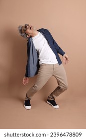 Asian energetic senior man with glasses posing  isolated on the brown background with casual clothes. mature man, Asian man. Elderly,  studio shot. Multiple emotions. - Shutterstock ID 2396478809