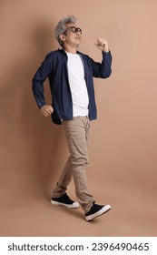 Asian energetic senior man with glasses posing  isolated on the brown background with casual clothes. mature man, Asian man. Elderly,  studio shot. Multiple emotions. - Shutterstock ID 2396490465