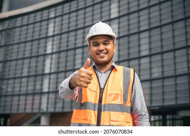Asian engineer handsome man or architect looking camera with white safety helmet in construction site. Standing at modern building construction. Worker asian man showing thumbs up.