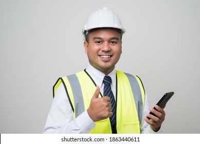 Asian engineer holding mobile phone and showing thumbs up. Worker wearing hard hat using smartphone standing on isolated white background.