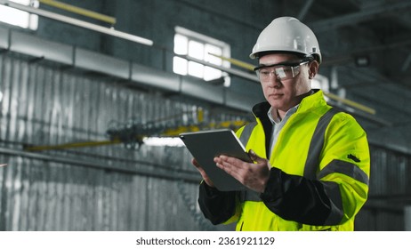 Asian engineer looks at screen of tablet. Worker wearing yellow vest, glasses and hard hat. Male stands on background of workflow. Man at workplace. - Powered by Shutterstock