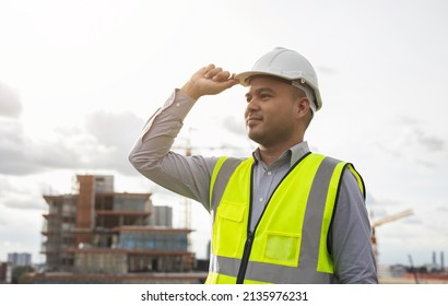 Asian Engineer Man Or Architect Looking Forward With White Safety Helmet In City Construction Site . Standing On Rooftop Building Construction At Capital.