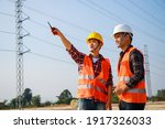 Asian engineer manager and foreman or leader discussion and pointing to construction site project on workplace and High voltage power line pylon in the background. Teamwork, Leadership concept.