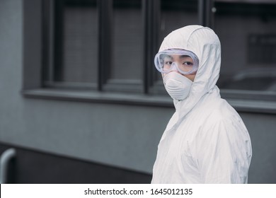 Asian Epidemiologist In Hazmat Suit And Respirator Mask Looking At Camera While Standing On Street