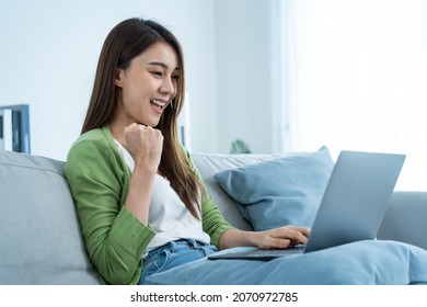 Asian excited young woman winner feeling happy after looks at laptop. Attractive girl student sitting on sofa in living room and found out good news on computer then celebrate online success in house.