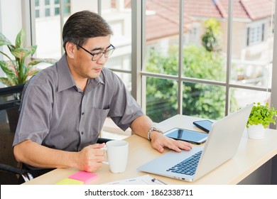 Asian Executive Senior Businessman Sitting On Desk Office With His Office, The Confident Middle Aged Handsome Man Using Laptop Computer At Workplace Home Office And Drinking A Cup Of Coffee