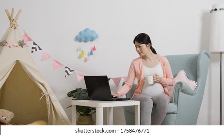 Asian Expectant Mother Shopping Online For Baby Items On Laptop With Coffee In Playroom. Japanese Female Worker Contemplating While Doing Project With Notebook. Technology And Work From Home Concept