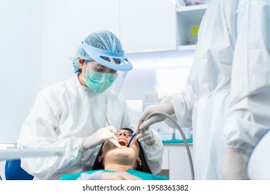 Asian Expert Dentist Doctor Wear PPE And Mask Examine Patient's Teeth At Dental Clinic. Dental Assistant Use Suction To Keep Young Girl's Mouth Dry Due Treatment Procedures During Coronavirus Pandemic