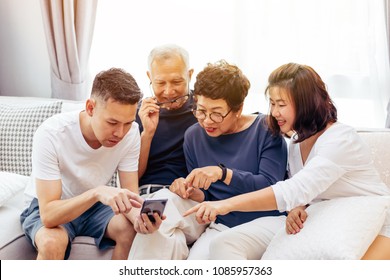 Asian Family With Adult Children And Senior Parents Using A Mobile Phone And Relaxing On A Sofa At Home Together