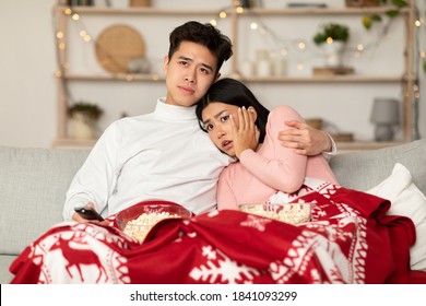 Asian Family Couple Watching Television And Horror Movie On TV Sitting On Couch Covered With Blanket At Home, Celebrating Christmas Together. Chinese Spouses Watch Scary Movies During Winter Holidays