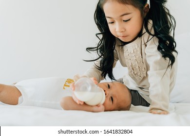 Asian Family Of Cute Little Sister Feeding Bottle Of Milk To Newborn Baby Boy Brother. Toddler Kid And New Sibling Relax In A White Bedroom At Home