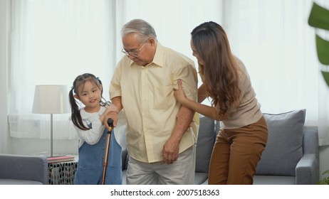 Asian Family, Daughter And Granddaughter Take Care Support Grandfather Who Is Suffering From Knee Pain Got Walking Outside To Take A Walk And Grandpa With A Stick, Elderly Health Care Concept