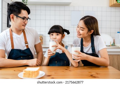 Asian  family enjoying breakfast at cozy kitchen, little girl daughter sitting on table, drinking milk with smiling father and mother in morning. Happy family in kitchen.