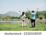 Asian family father mother and daughter stand front lake with raised hands sunny day in park, lifestyle parents and child kid enjoying outdoors together in nature on summer travel, Family relaxation