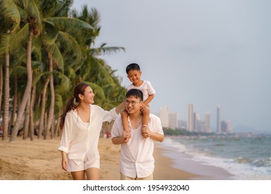 Asian family with fathers carrying his son on back and mother walking togerther along a beachfront beach with coconut trees while on vacation in the summer in Thailand.