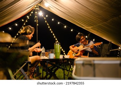 Asian Family And Friends Vacation Camp, Dinner And Relaxing Guitar Play Under Tarp Or Flysheet, Outdoor Camping On The Courtyard With Warm Light At Night Time.