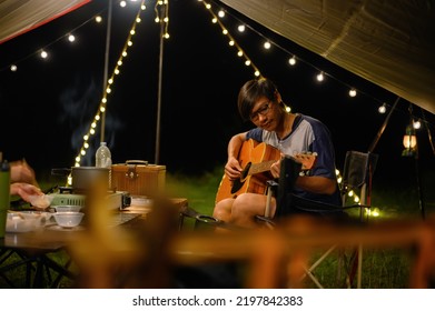 Asian Family And Friends Vacation Camp, Dinner And Relaxing Guitar Play Under Tarp Or Flysheet, Outdoor Camping On The Courtyard With Warm Light At Night Time.