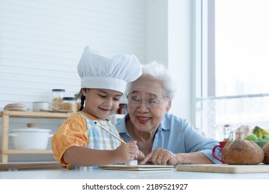 Asian Family Grandmother And Grandchild Have Fun Cooking At Home Kitchen Together, Little Cute Granddaughter With Chef Hat And Apron Is Writing Down Recipe And How To Make Bread Or Cookie From Grandma